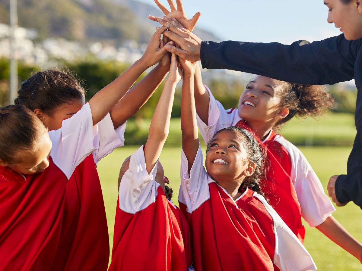 A group of children in red jerseys and a coach raise their hands in a huddle on a sports field, showing teamwork and excitement together.
