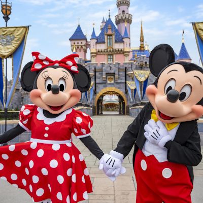 Mickey and Minnie Mouse stand with a child in a polka-dot dress; a castle backdrop and festive crowns create a magical, playful scene.