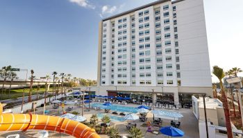 A large hotel with a tall white facade overlooks a pool areaWith slides, lounge chairs, and blue umbrellas around the pool in front of the building, sunny day.