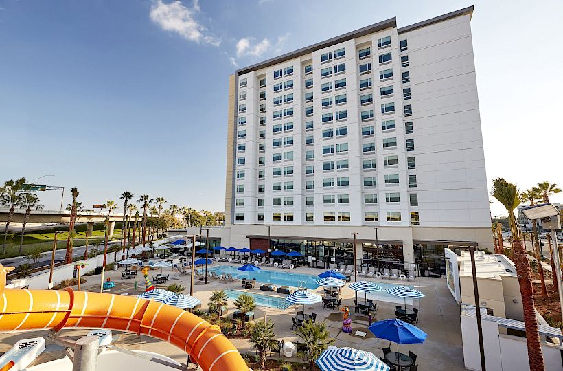 A large hotel with a tall white facade overlooks a pool areaWith slides, lounge chairs, and blue umbrellas around the pool in front of the building, sunny day.