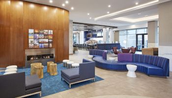 A modern hotel lobby with curved blue seating, wooden accent wall, small stools, a blue rug, and a bright, airy space with lots of chairs and tables.