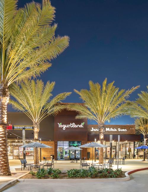 A shopping plaza at night with palm trees, outdoor seating, and storefronts; a modern, lit-up strip mall vibe.