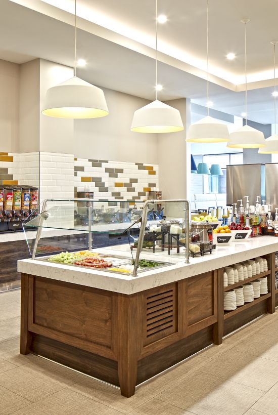 A modern cafeteria or buffet setup with a long central serving island, warm lighting, and a self-serve section in the background.