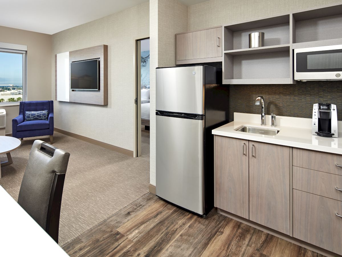 A compact modern kitchen with a stainless fridge, microwave, sink, and cabinets, adjacent to a living area with a TV and seating, ending here.