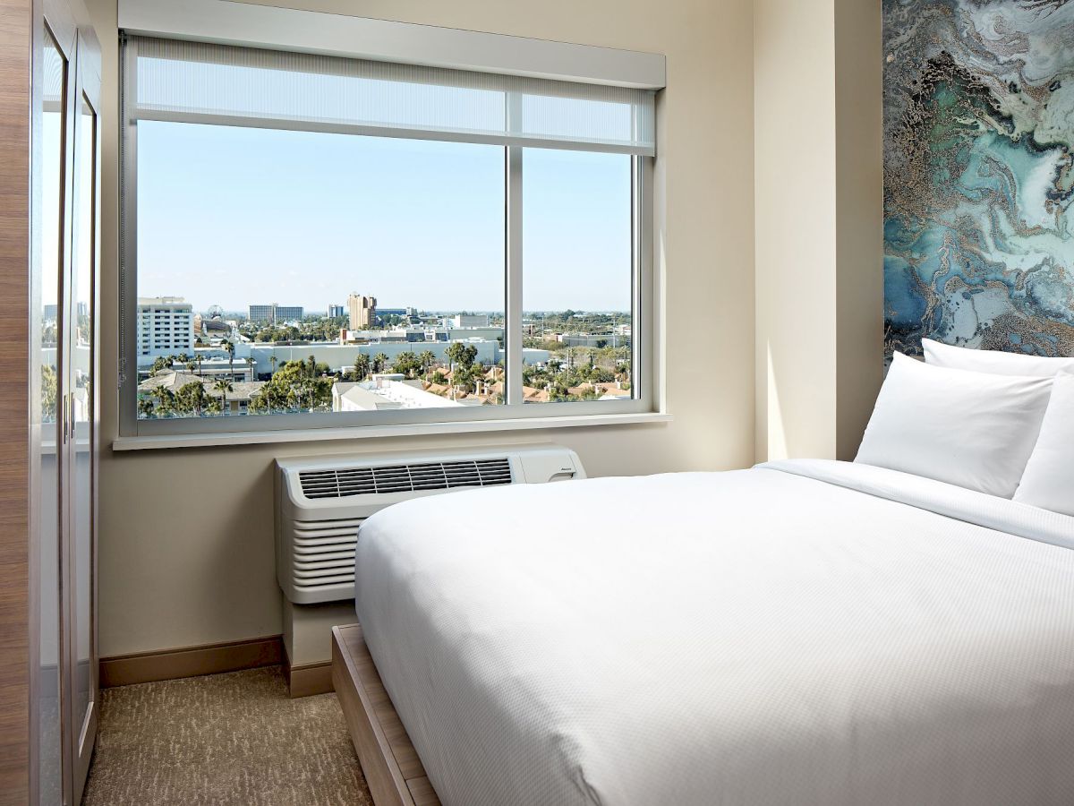 A clean hotel room with a large window view, a white bed, light decor, and an air conditioner unit below the window, overlooking a cityscape.
